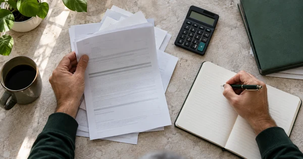 Des mains examinent des documents et prennent des notes à côté d’une calculatrice et d’un dossier vert.